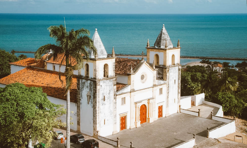 Igreja com torres brancas cercada por coqueiros, com o mar ao fundo